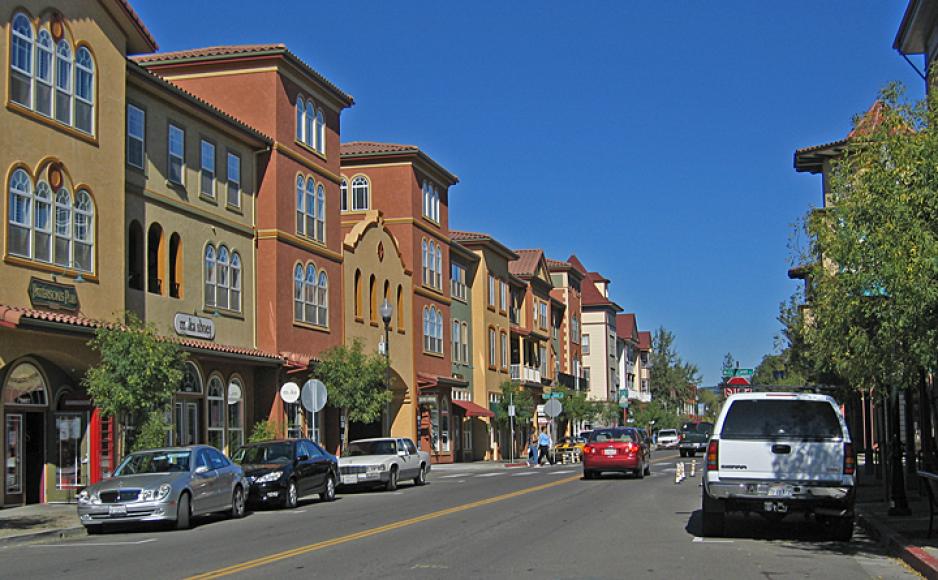 Photo of a small mixed use street lined by shops with residences above, street trees, wide sidewalks, pedestrians crossing.